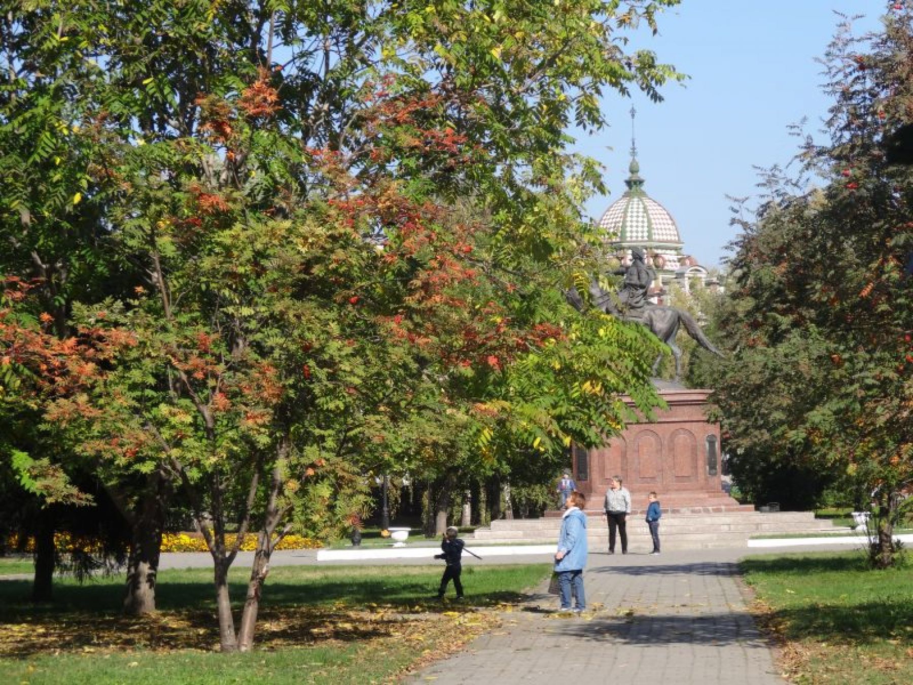 Памятник стоит посреди парка, в окружении традиционных для юга Сибири хвойных и лиственных пород деревьев и кустарников 